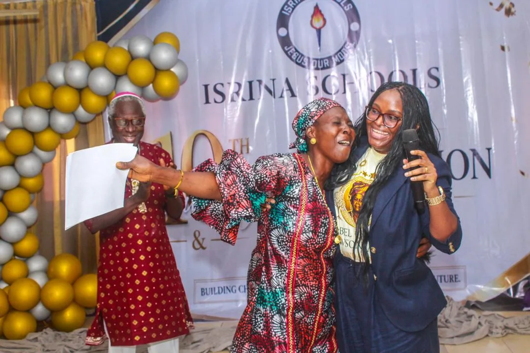 Two women celebrating on stage, one holding a sheet of paper, with an ISRINA Schools backdrop